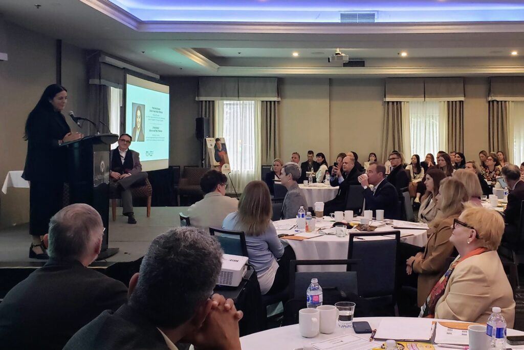 Photo of a conference room filled with people seated in groups at round tables. On a stage on the left, the Minister of Tourism speaks at a podium.