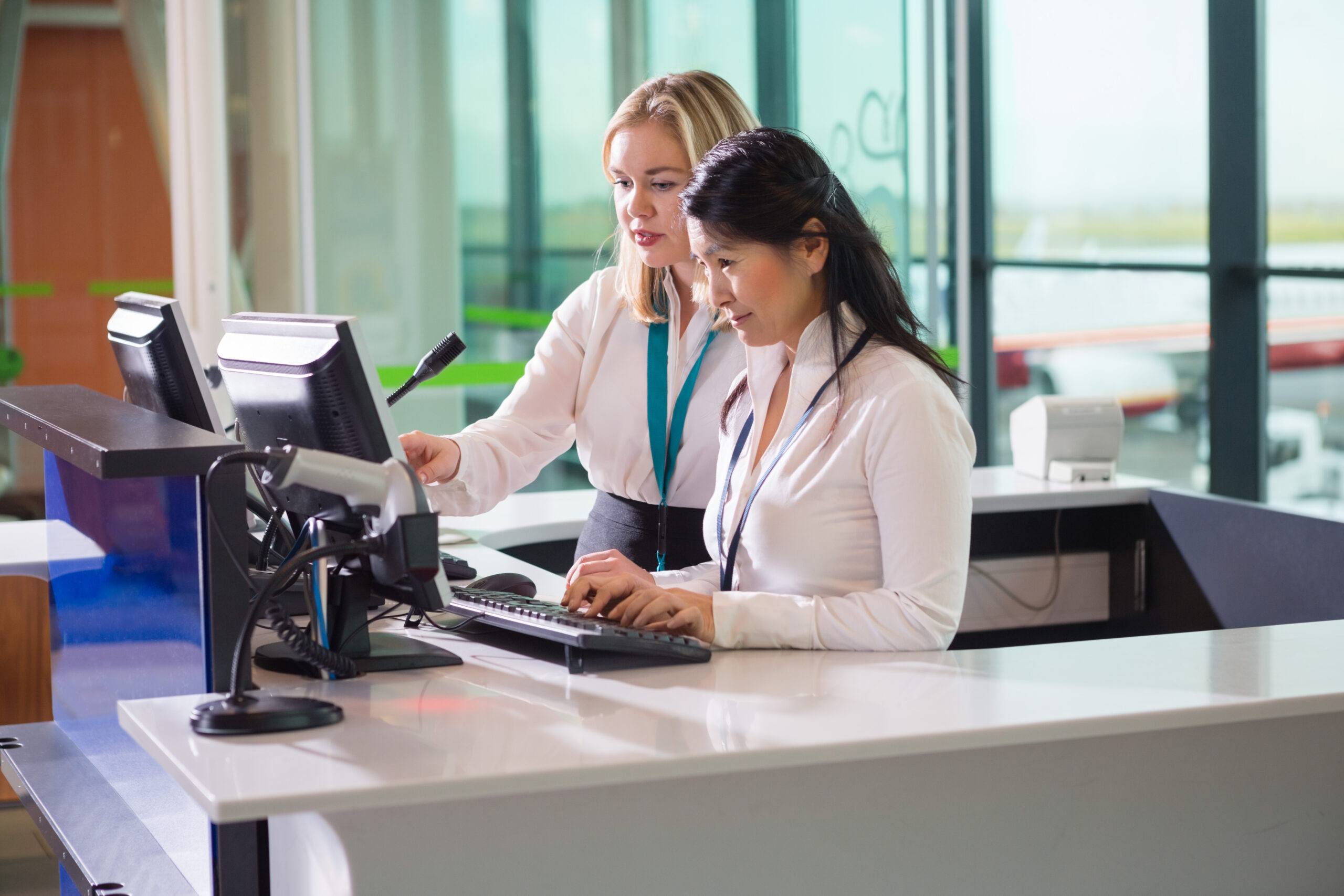 Two female airline workers at the gate desk. One worker is showing the other how to operate the computer.