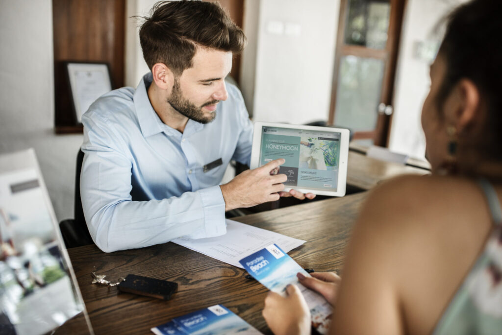 Male travel agent pointing to information on a tablet to a female on the other side of the table.