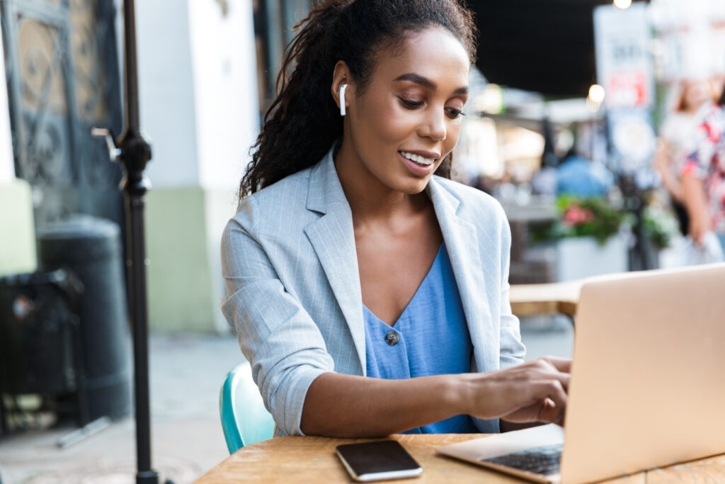Female sitting at a table outdoors with her laptop. She is entering on her keyboard, wearing earbuds in her ear.
