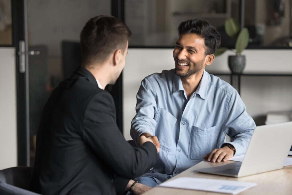 Cheerful Multiethnic Male Business Partners Shaking Hands At Workplace Table,