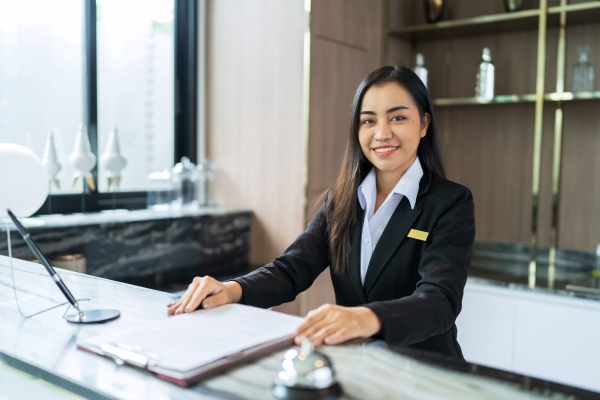 A female hotel front desk agent smiling at the camera. She is standing behind the counter with a clipboard in front of her on the counter.