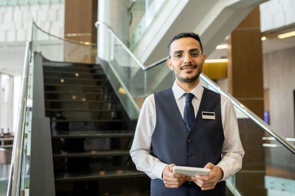 A hotel staff member stands in front of a sunlight staircase, holding a tablet and smiling at the viewer.
