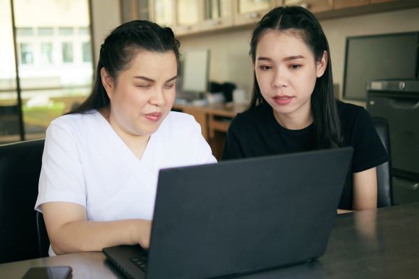 Two women are sitting at a desk in a workplace setting. One, who has a visual disability, is using a laptop while the other is speaking to her and looking at the monitor.