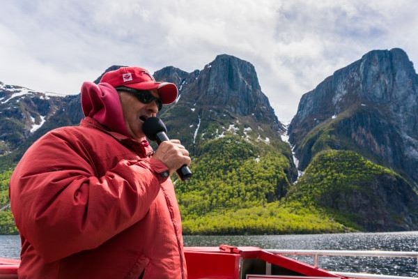 A tour guide on a boat speaks into a microphone. Mountains and a waterfall in the background.