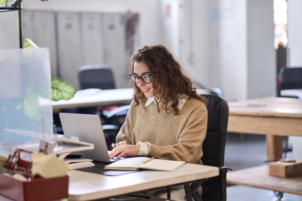 A woman sits at a desk in an open plan office. She is working on a laptop.