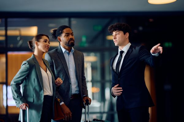 A male hotel worker in a suit points off camera, giving directions to a male and female in business attire, who are each pushing a suitcase.
