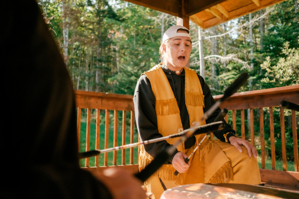 An Indigenous individual sings as they drum. Several other individuals are also drumming, but are set outside the frame of the image. The group is on a veranda or porch, and there is a forested area beyond the lawn behind them.