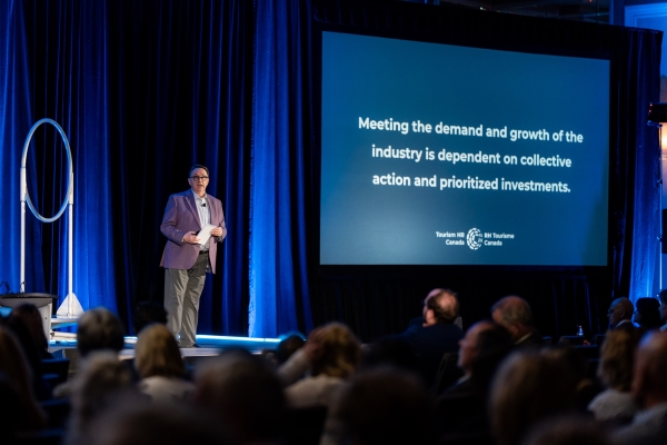 A speaker is on a stage, presenting to a group at a conference. A screen in the background displays the text: Meeting the demand and growth of the industry is dependent on collective action and prioritized investments.