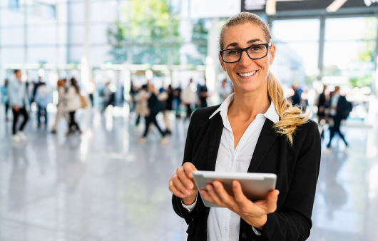 Lady smiling at the camera holding an ipad standing in a busy work office.