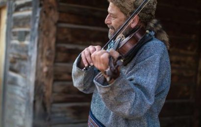 Two heritage performers playing the violin outside of a cabin.