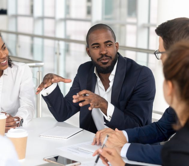 Male office worker in discussion with a group, all sat around a table. There are papers and coffee cups on the table.