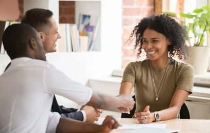 Female sat down facing two male in an office setting. She is shaking the hand of one of the opposing males, smiling.