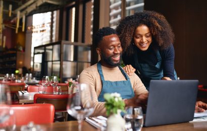 Male restaurant worker sitting at a table with an open laptop. There is a female restaurant worker standing beside him, crouching down to view the laptop screen. They are both smiling.
