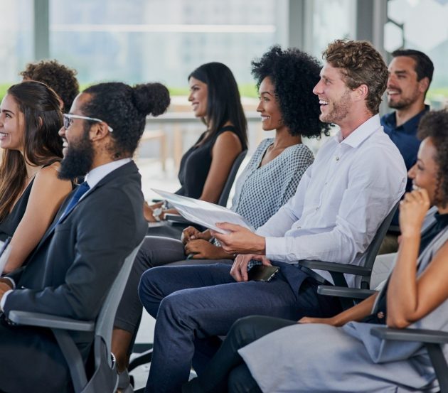 shutterstock_2485558527 A working group of people sitting in chairs in a large room. They are all facing the same direction, smiling. Some are holding notepads.