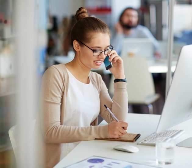 Female sitting at a desk in front of a desktop. She is on the phone, writing notes on a notepad.