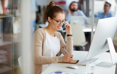 Female sitting at a desk in front of a desktop. She is on the phone, writing notes on a notepad.