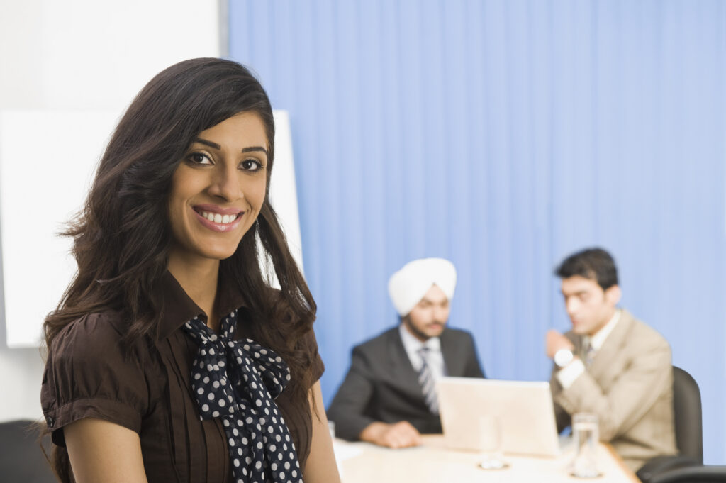 Female of middle eastern nationality looking at the camera, smiling. There are two men behind her sitting at a table looking at a laptop.