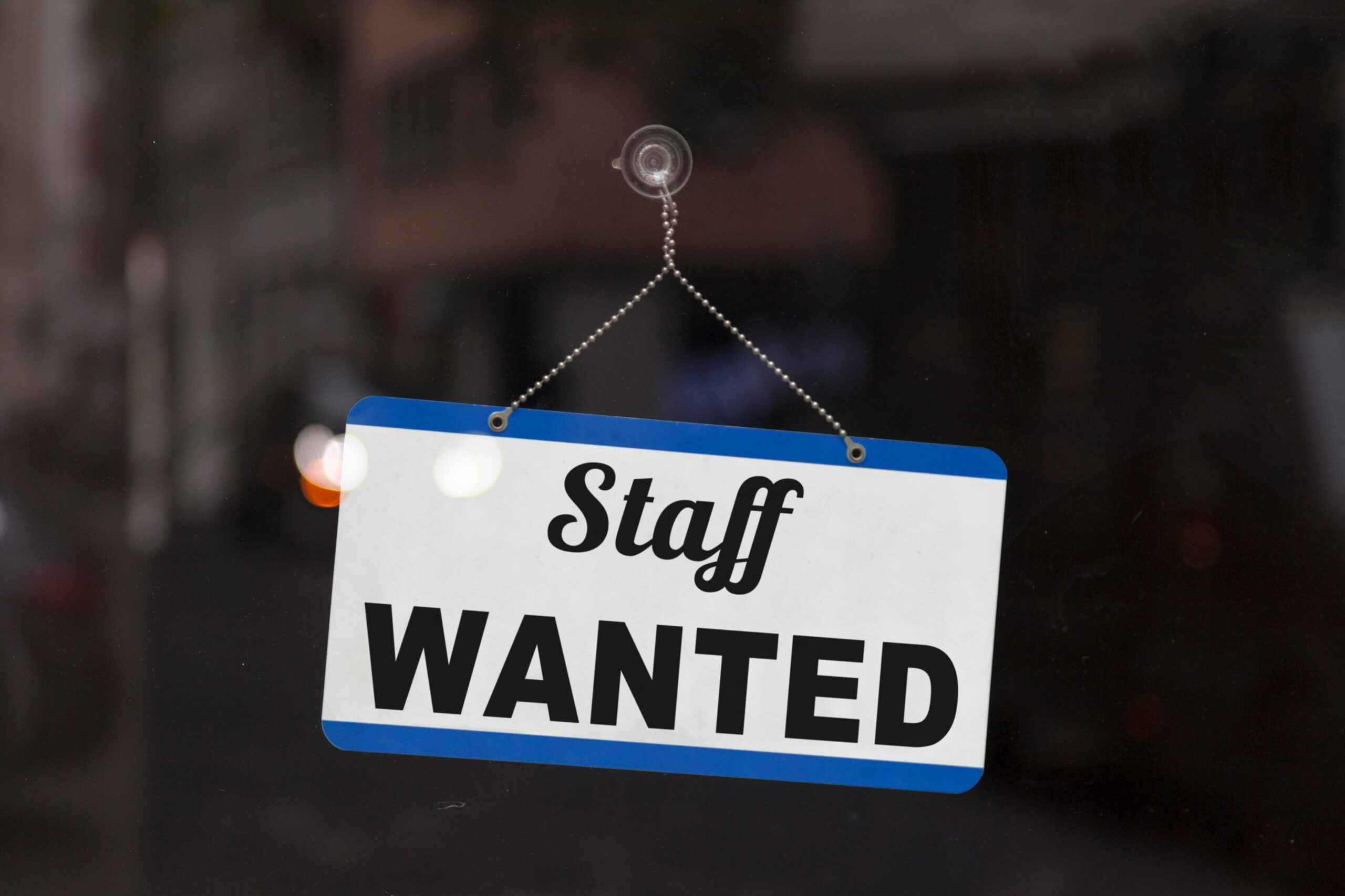 Hanging sign on a glass door that says "Staff Wanted"