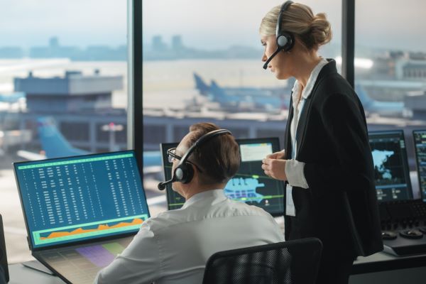 Image of two air traffic controllers consulting a computer screen, in the air traffic control tower overlooking the airport.