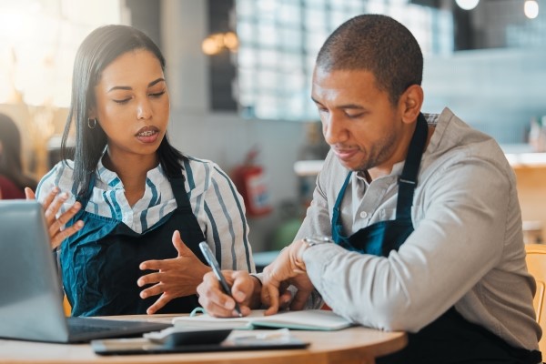 Two restaurant managers sit at a table discussing paperwork. One speaks while the other writes in a notebook. A laptop is on the table in front of them.