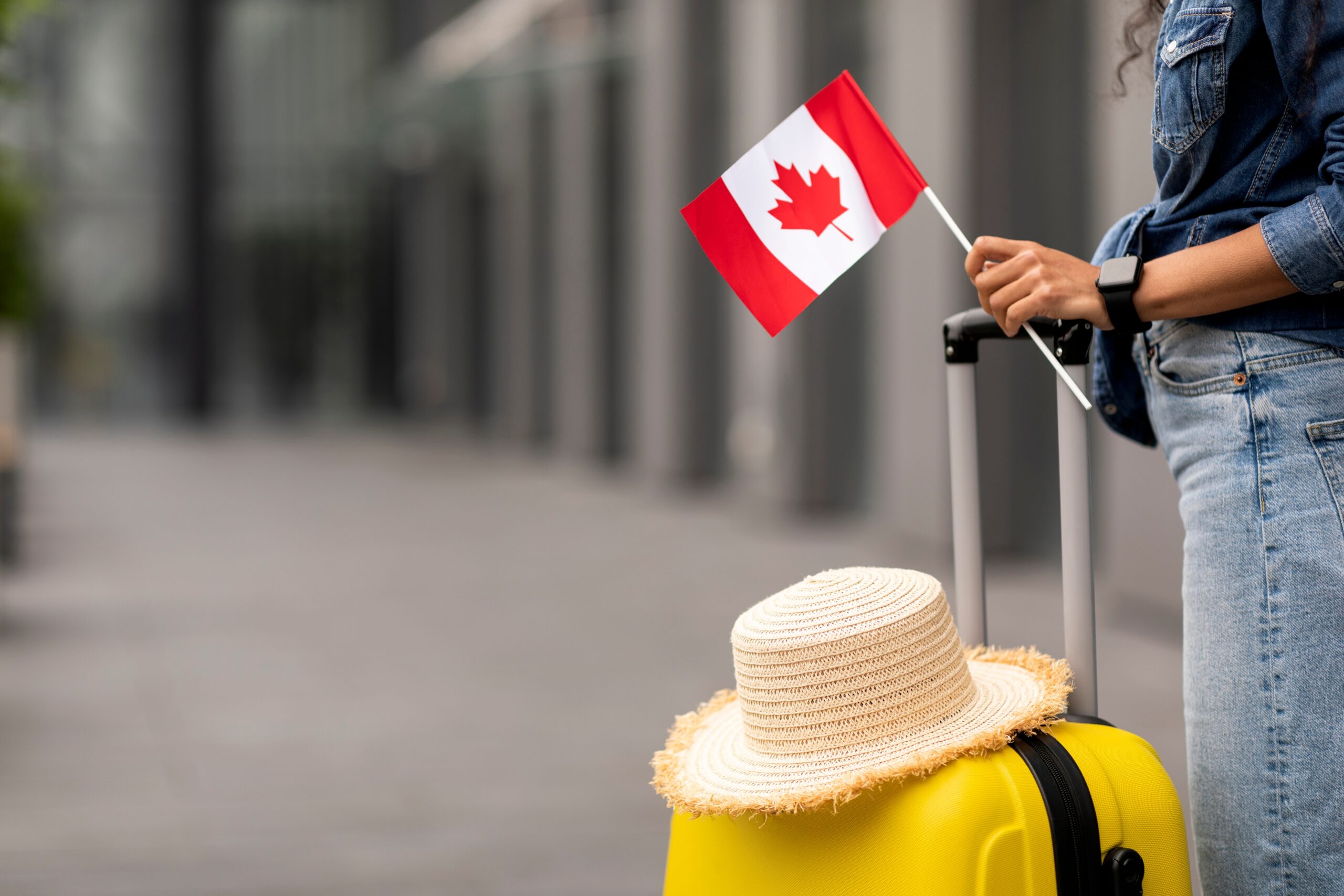 A person standing by their yellow suitcase. There is a straw hat resting on the top of the suitcase and the person is holding a small Canada flag.