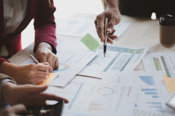 A collection of printed materials with graphs and charts are on a table, taking up most of the frame. One person's hand points at a document with a pen, while another person's hands are poised with a pencil above a sticky note. A third person's hand points at another document.