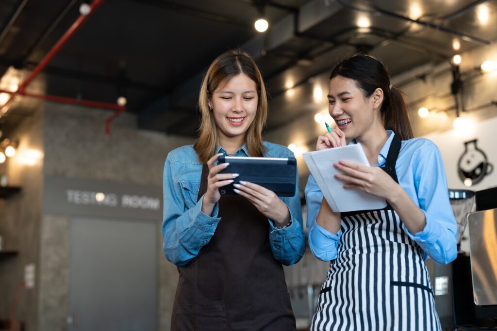 Two women working in a restaurant, one is holding an iPad and the other is writing notes. They are both standing and smiling.