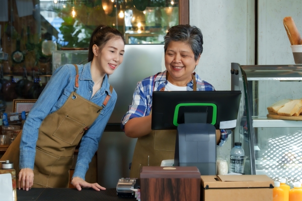 Two women are working in a café. The older woman is showing the younger woman how to use the cash register.