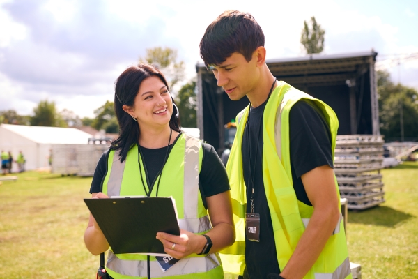 Two event staff members consult a clipboard at an outdoor festival venue. A stage, a large white tent, and sound equipment are in the background.