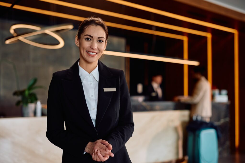 Female front desk agent smiling at the camera. She is standing in the lobby of the hotel with the front desk behind her. She is wearing a black blazer and collared white shirt.