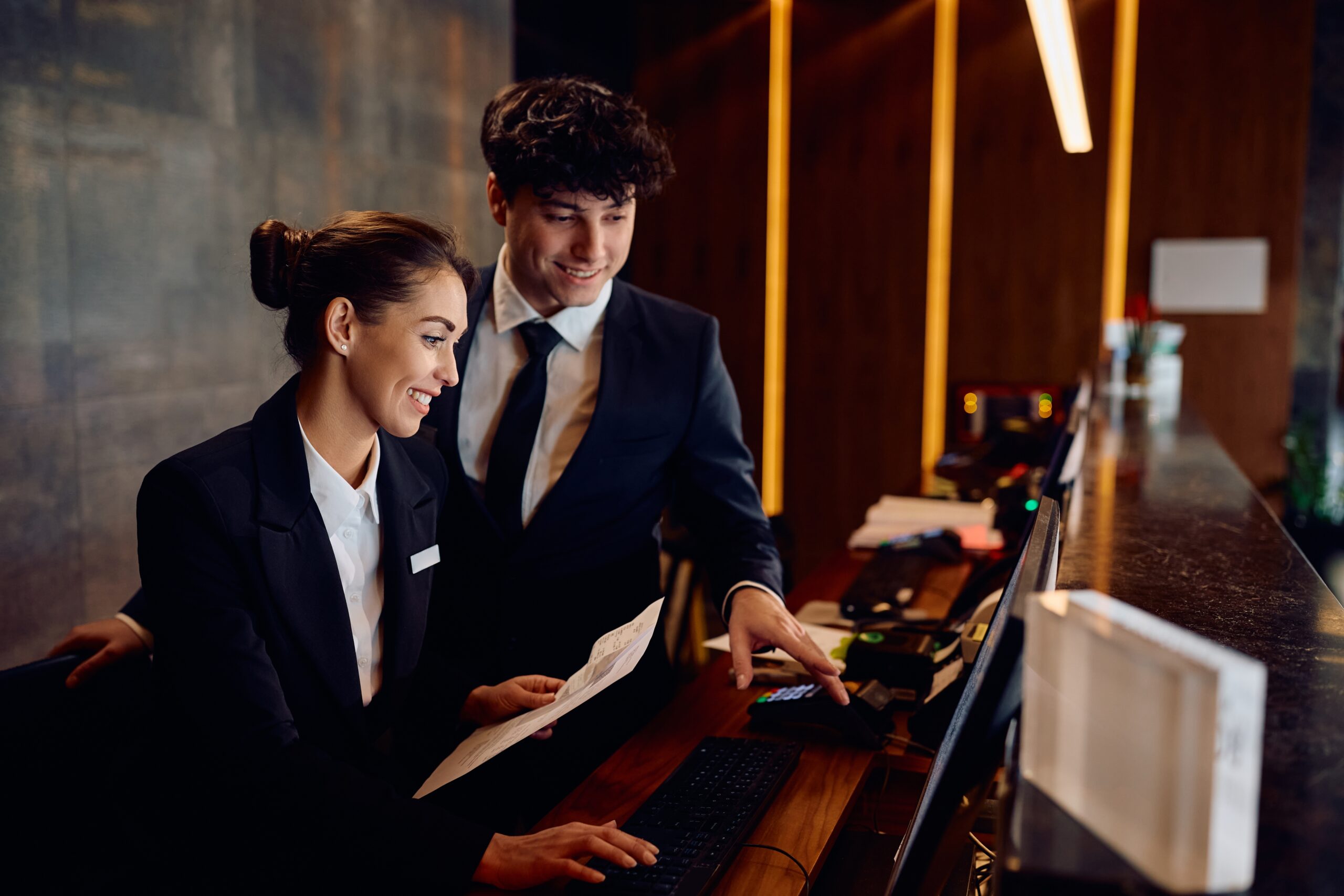 Two hotel front desk agents behind the desk. One of pointing to the computer screen, and the other is holding paper while inputting an entry.