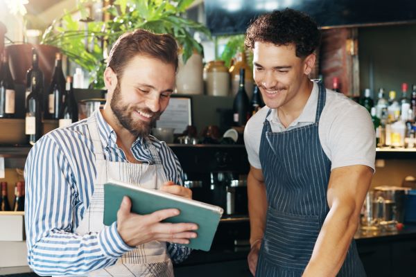 A restaurant worker shows a younger colleague how to do a task on a tablet.
