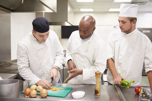 A cooking instructor provides a student with guidance on slicing vegetables, in a commercial kitchen.